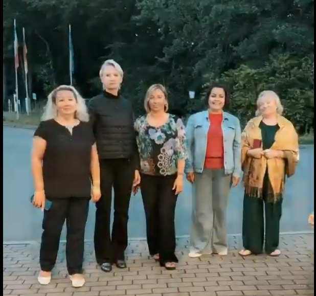 Gruppe von fünf Frauen steht draußen auf einem gepflasterten Weg vor Bäumen und Grünfläche